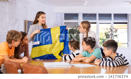 Joyful female teacher discussing with preteens students state flag of Sweden in auditory of middle school Joyful female teacher discussing with preteens students state flag of Sweden in auditory of middle school 141736374