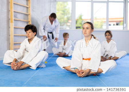 Children in white kimono sits in butterfly pose and practices stretching in sport gym Children in white kimono sits in butterfly pose and practices stretching in sport gym 141736391
