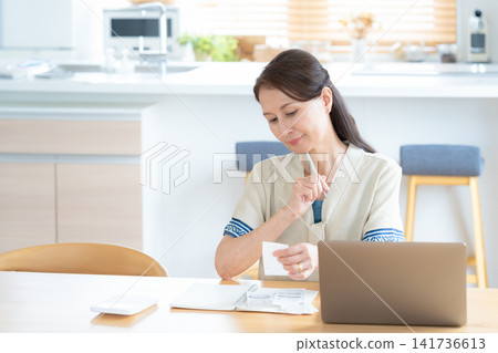 A senior woman keeping track of household expenses in the living room. A senior woman keeping track of household expenses in the living room. 141736613