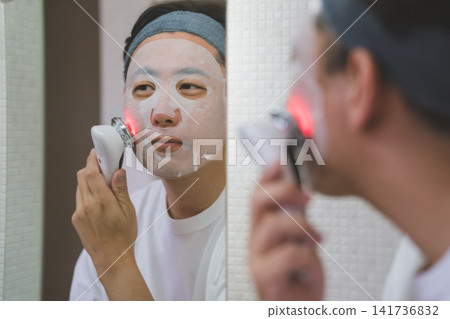 一位 40 歲男子的護膚流程:對著鏡子敷面膜,同時使用 LED 臉部美容儀撫平臉頰。 一位 40 歲男子的護膚流程:對著鏡子敷面膜,同時使用 LED 臉部美容儀撫平臉頰。 141736832