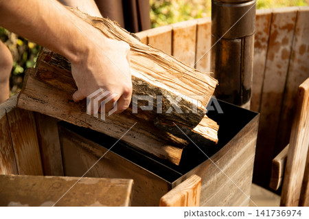 Hands place logs into wood-burning stove in garden. Outdoor cooking and warmth concept Hands place logs into wood-burning stove in garden. Outdoor cooking and warmth concept 141736974