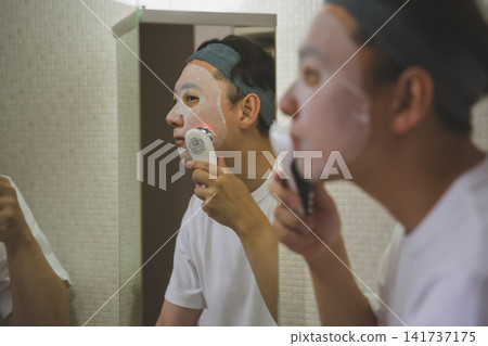 一位 40 歲男子的護膚流程:對著鏡子敷面膜,同時用紅色 LED 臉部儀按摩臉頰。 一位 40 歲男子的護膚流程:對著鏡子敷面膜,同時用紅色 LED 臉部儀按摩臉頰。 141737175