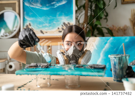 Serious woman artist wearing gloves and respirator working with epoxy resin in creative workshop. 141737482