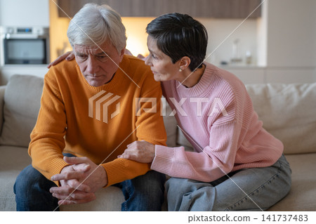 Senior couple sitting on sofa at home, emphatic elderly woman comforts upset overwhelmed husband. Senior couple sitting on sofa at home, emphatic elderly woman comforts upset overwhelmed husband. 141737483