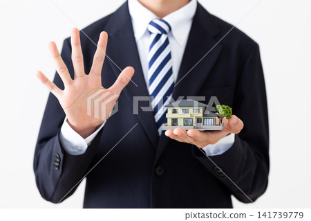 A young businessman in a suit holds up five fingers while holding up a model of a house. 141739779