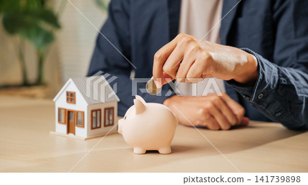 A businessman hand places a coin into a piggy bank beside a house model, symbolizing property savings, financial planning, and investment strategies for home buying A businessman hand places a coin into a piggy bank beside a house model, symbolizing property savings, financial planning, and investment strategies for home buying 141739898