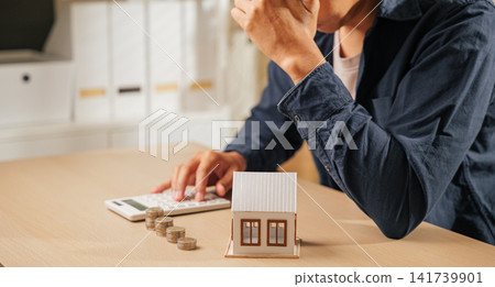Stressed person holding a calculator, with stacks of coins and a house model nearby. Reflects financial planning challenges, tax concerns, and property calculations causing worry Stressed person holding a calculator, with stacks of coins and a house model nearby. Reflects financial planning challenges, tax concerns, and property calculations causing worry 141739901