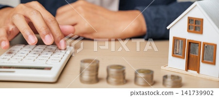 A person presses a calculator next to a house model and coin stacks, analyzing tax, property value, and mortgage payments. Highlights real estate profit, cash savings A person presses a calculator next to a house model and coin stacks, analyzing tax, property value, and mortgage payments. Highlights real estate profit, cash savings 141739902