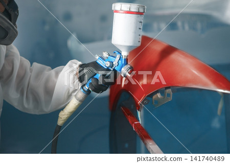 Car paint worker spraying red paint to car body element using spray gun in vehicle workshop chamber. Complex restoration of automobile body. Car paint worker spraying red paint to car body element using spray gun in vehicle workshop chamber. Complex restoration of automobile body. 141740489