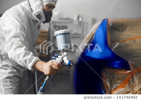 Automobile repairman painter in mask and protective workwear painting car body in paint chamber. Automobile repairman painter in mask and protective workwear painting car body in paint chamber. 141740492
