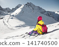 Woman in Bright Winter Attire Sits on Snowy Slope Admiring Majestic Mountain View on Sunny Day 141740500