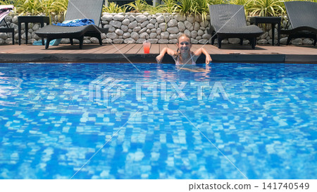 Teenage girl relaxing and floating in a blue resort swimming pool with a strawberry cocktail in a glass placed on the pool deck nearby. Pool relaxation concept Teenage girl relaxing and floating in a blue resort swimming pool with a strawberry cocktail in a glass placed on the pool deck nearby. Pool relaxation concept 141740549