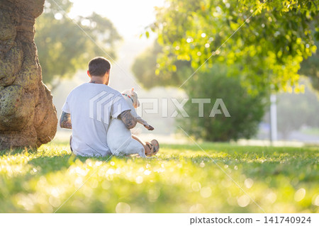 Old bull terrier sitting on the lap of a tattooed male owner in a green park, sunlight filtering through trees creating a serene outdoor atmosphere Old bull terrier sitting on the lap of a tattooed male owner in a green park, sunlight filtering through trees creating a serene outdoor atmosphere 141740924