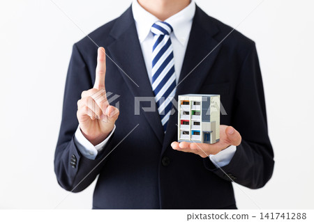 A young businessman in a suit holds up a model of an apartment building and raises his index finger. A young businessman in a suit holds up a model of an apartment building and raises his index finger. 141741288