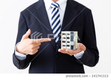 A young businessman in a suit points to a model of an apartment building. A young businessman in a suit points to a model of an apartment building. 141741313
