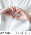 Close up of a child forming a heart shape with both hands against a white shirt, expressing love, kindness, affection, and emotional connection through a simple hand gesture. 141741635