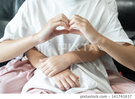 An older sister forms a heart with her hands as her younger sister embraces her from behind, symbolizing care, unity, and emotional connection. An older sister forms a heart with her hands as her younger sister embraces her from behind, symbolizing care, unity, and emotional connection. 141741637