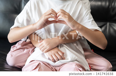 A younger sister hugs her older sister from behind while the older sister makes a heart sign, representing unconditional love and emotional support. A younger sister hugs her older sister from behind while the older sister makes a heart sign, representing unconditional love and emotional support. 141741638