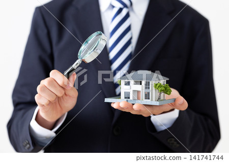 A young businessman in a suit holding a model of a detached house and a magnifying glass. A young businessman in a suit holding a model of a detached house and a magnifying glass. 141741744