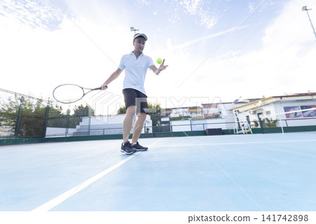 Tennis player practices serve on a blue court in the late afternoon at a sports facility Tennis player practices serve on a blue court in the late afternoon at a sports facility 141742898