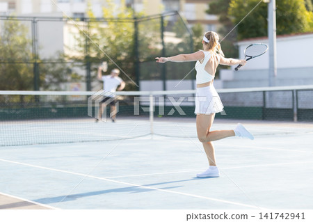 Woman plays tennis on a court while a man stands in the background at a sunny day in a sports facility Woman plays tennis on a court while a man stands in the background at a sunny day in a sports facility 141742941