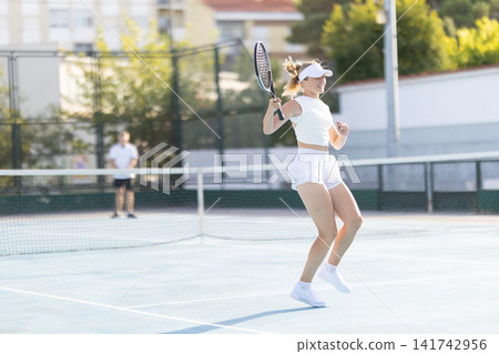 Young woman plays tennis on a court during daytime in a warm climate with a man in the background Young woman plays tennis on a court during daytime in a warm climate with a man in the background 141742956