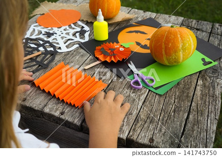 Close up of a little girl sitting at a rustic wooden table outdoors with colorful Halloween craft materials 141743750