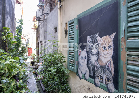 Colorful mural featuring four playful cats painted on a wall in a narrow alley, surrounded by lush greenery and vibrant plants, creating a whimsical urban atmosphere 141743973