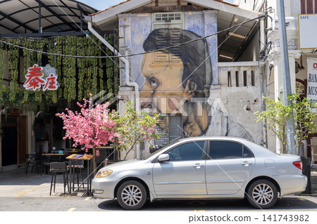 Penang, Malaysia - May 17, 2024: Street art mural of a man on building wall beside a silver car parked on the street with blooming pink flowers and vibrant cafe atmosphere Penang, Malaysia - May 17, 2024: Street art mural of a man on building wall beside a silver car parked on the street with blooming pink flowers and vibrant cafe atmosphere 141743982