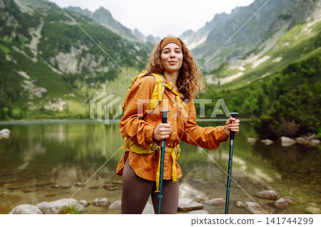 Female traveler with hiking backpack poses against backdrop of mountain lake. Adventure concept. Female traveler with hiking backpack poses against backdrop of mountain lake. Adventure concept. 141744299