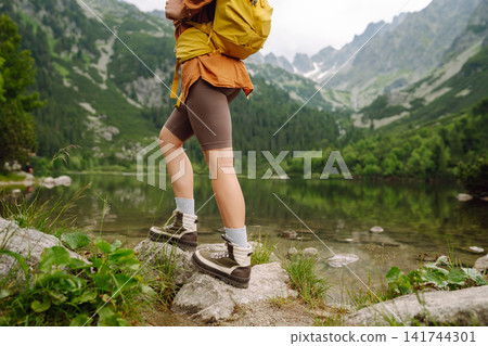 Female traveler with hiking backpack poses against backdrop of mountain lake. Adventure concept. Female traveler with hiking backpack poses against backdrop of mountain lake. Adventure concept. 141744301