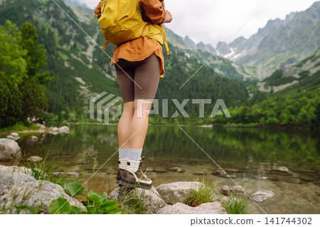 Female traveler with hiking backpack poses against backdrop of mountain lake. Adventure concept. Female traveler with hiking backpack poses against backdrop of mountain lake. Adventure concept. 141744302