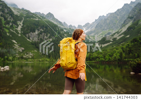 Female traveler with hiking backpack poses against backdrop of mountain lake. Adventure concept. 141744303