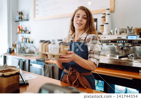 The young barista owner behind the bar gives out to-go orders. Business concept. Drinks to go. The young barista owner behind the bar gives out to-go orders. Business concept. Drinks to go. 141744354