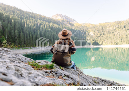 Back view young woman with backpack looking the view of the mountain lake in sunny weather. Back view young woman with backpack looking the view of the mountain lake in sunny weather. 141744561