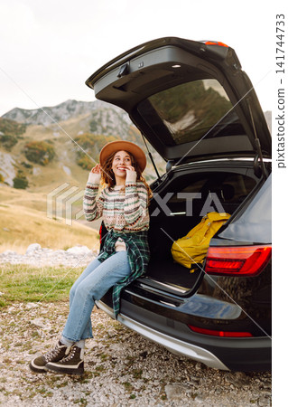 A traveler woman with curly hair and a hat smiles as she relaxes at the back of her black SUV A traveler woman with curly hair and a hat smiles as she relaxes at the back of her black SUV 141744733