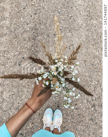 Hand holding wildflower bouquet with chamomile and dry grass, top view on pavement background, casual summer lifestyle Hand holding wildflower bouquet with chamomile and dry grass, top view on pavement background, casual summer lifestyle 141744897