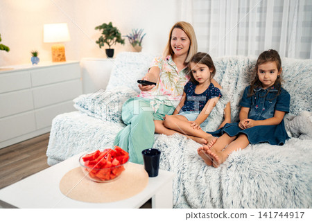 Mother and daughters watching TV relaxing together on couch Mother and daughters watching TV relaxing together on couch 141744917