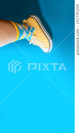 Leg wearing a yellow sneaker and colorful patterned socks against a vibrant bright blue background, modern fashion and youth lifestyle concept Leg wearing a yellow sneaker and colorful patterned socks against a vibrant bright blue background, modern fashion and youth lifestyle concept 141745204