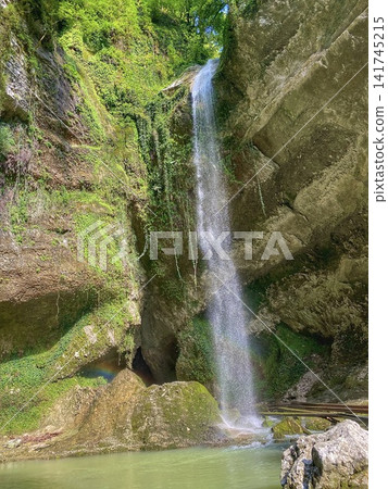 Waterfall flowing into clear pool in green forest with small rainbow and sunlight on rocks Waterfall flowing into clear pool in green forest with small rainbow and sunlight on rocks 141745215