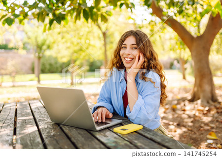 Attractive woman working or studying at laptop as sitting in city park. Freelancer working in park. 141745254
