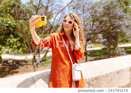 Young woman enjoys sunny day by taking selfie in vibrant park filled with blooming flowers. Blogging Young woman enjoys sunny day by taking selfie in vibrant park filled with blooming flowers. Blogging 141745274