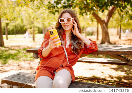 Young woman sits on bench in park, using smartphone while dressed casually. Travel, blogging concept Young woman sits on bench in park, using smartphone while dressed casually. Travel, blogging concept 141745276