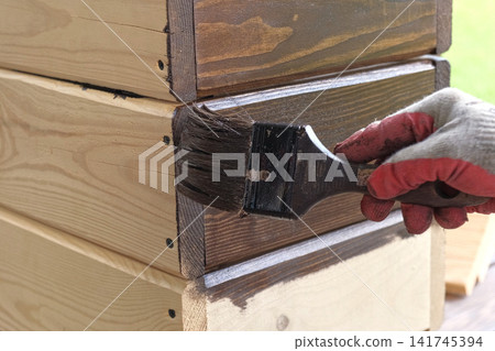 Hand in glove painting wooden box with brown oil stain brush. Worker applying protective varnish on pine timber planks surface, DIY woodwork. Wood staining process. Backyard Woodworking, Maintenance. 141745394
