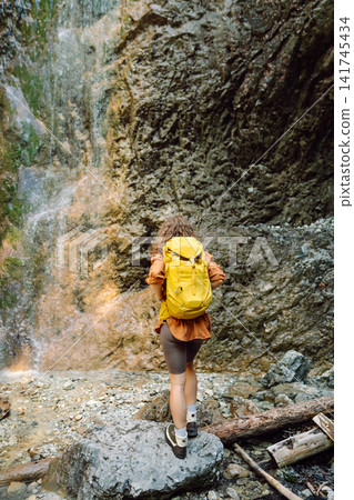 Smiling female dressed in hiking clothes, enjoys a waterfall near a mountain river. Travel, trekking Smiling female dressed in hiking clothes, enjoys a waterfall near a mountain river. Travel, trekking 141745434