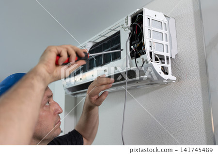 Technician repairing air conditioner with screwdriver. Professional maintenance of cooling system. Indoor climate control device repair service, daylight, low angle shot. Technician repairing air conditioner with screwdriver. Professional maintenance of cooling system. Indoor climate control device repair service, daylight, low angle shot. 141745489