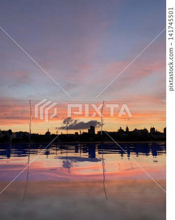 Colorful sunset sky reflected in water with city skyline silhouette and soft ripples in foreground Colorful sunset sky reflected in water with city skyline silhouette and soft ripples in foreground 141745501
