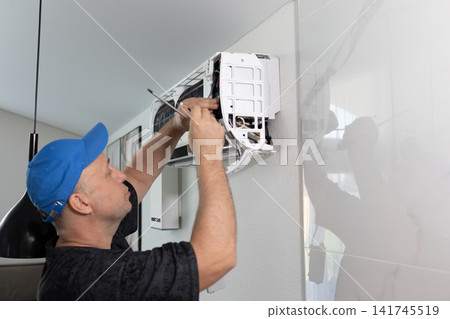 Technician man repairing air conditioning unit on wall. Professional maintenance worker using screwdriver. Climate control system service at home. Daytime soft lighting side view shot. Technician man repairing air conditioning unit on wall. Professional maintenance worker using screwdriver. Climate control system service at home. Daytime soft lighting side view shot. 141745519