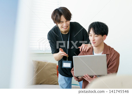 Two young Japanese men talking while looking at laptops in an office. Two young Japanese men talking while looking at laptops in an office. 141745546