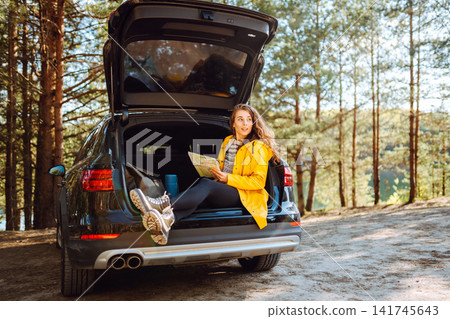 Woman searching location on map sitting on trunk of hatchback car outdoors. Road trip, travel. Woman searching location on map sitting on trunk of hatchback car outdoors. Road trip, travel. 141745643